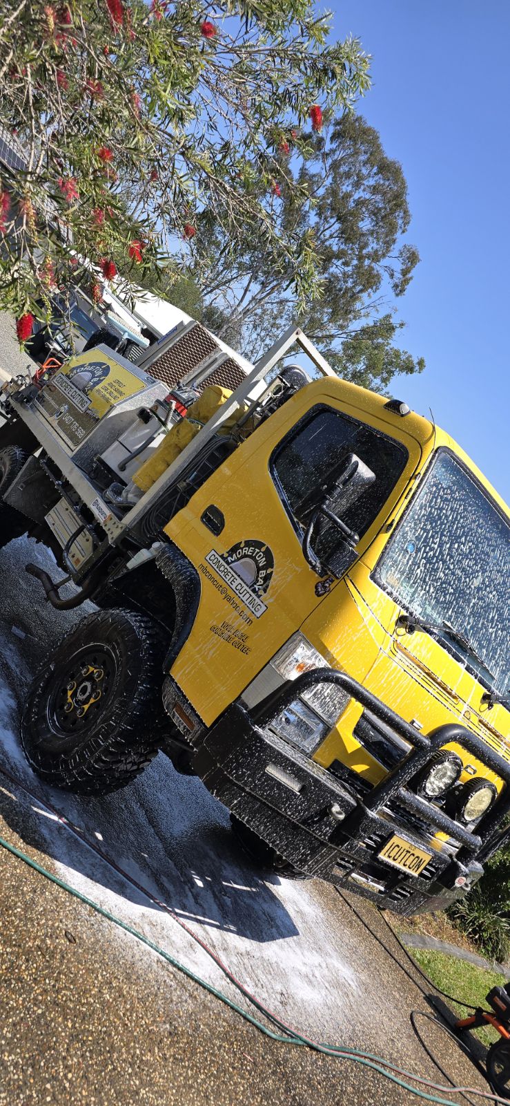 The image showcases a yellow concrete cutting truck being washed, with foam covering its surface, suggesting a maintenance routine. The truck is parked on a gravel surface surrounded by trees, indicating an outdoor setting. Its bright colour makes it stand out, while the foam hints at a thorough cleaning process essential for a concrete contractor. The clear blue sky in the background adds to the vividness of the scene, portraying a sunny day conducive for outdoor work.