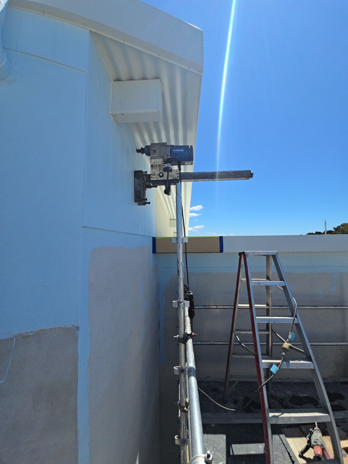 The image shows a construction setting featuring a ladder and an extended tool attached to the side of a building. The tool appears to be a concrete cutting saw, indicating a precise cutting process is underway, likely for installation or renovation purposes. This setup, complemented by the clear blue sky, suggests a well-maintained project site typical for concrete contractors. The equipment is positioned to work on the upper part of the wall, possibly preparing for a concrete feature or repairing an existing structure.