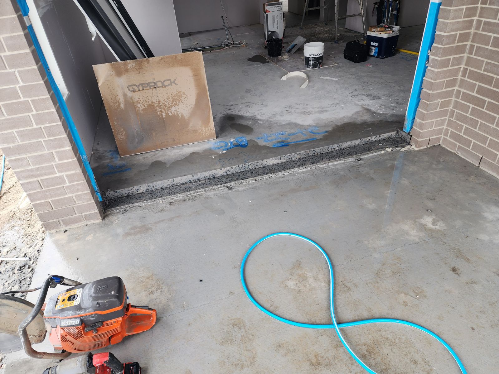 The image shows a construction site with a focus on a concrete floor that appears to have been cut or is in the process of being worked on. There are concrete cutting tools visible, including a chainsaw and a hose, indicating ongoing concrete work. In the background, there are construction materials and equipment, such as buckets and a piece of cardboard that reads 'OVERPACK'. The area appears to be prepped for further concrete installation or finishing work, with blue tape marking sections of the floor. The space looks open and under construction, suggesting it is part of a larger renovation or building project.
