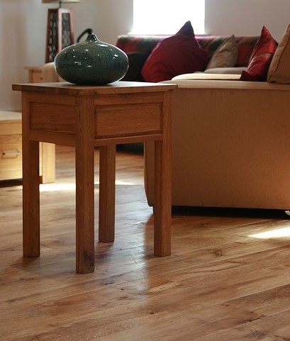 The image showcases a stylish and modern living space featuring high-quality wooden flooring. The light-coloured wooden floorboards enhance the room's brightness and warmth, providing a welcoming atmosphere. A small, wooden side table is positioned beside a plush sofa adorned with colourful cushions, indicating a cozy and inviting living area. In the centre of the table sits a green ceramic vase, adding an artistic touch to the decor. The overall design suggests a blend of comfort and elegance, highlighting the beauty of polished wood floors that AAA Floor Sanding, Concrete Polishing and Grinding might focus on in their projects.