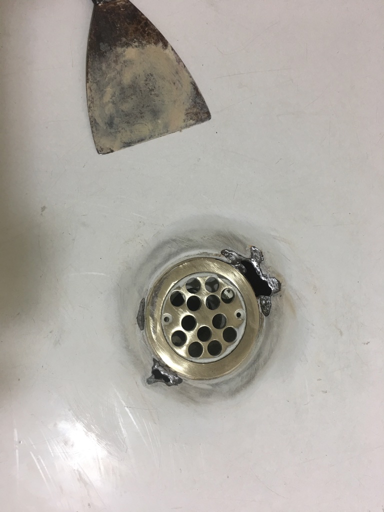 The image shows a close-up view of a bath with a visible drain. The drain appears to be partially disassembled, revealing some wear and damage around its edges. The surrounding area of the bath has a consistent surface, indicating it may be undergoing resurfacing or repair. A metal spatula is positioned above the drain, suggesting it is being used for scraping or applying a surface coating. This scene highlights the restoration process that Coastline Bath Resurfacing would typically undertake, where they remove old materials and prepare the bath for resurfacing to ensure a smooth, durable finish. This aligns with their services of enhancing and renewing bathroom fixtures for customers in Kyogle and nearby regions.