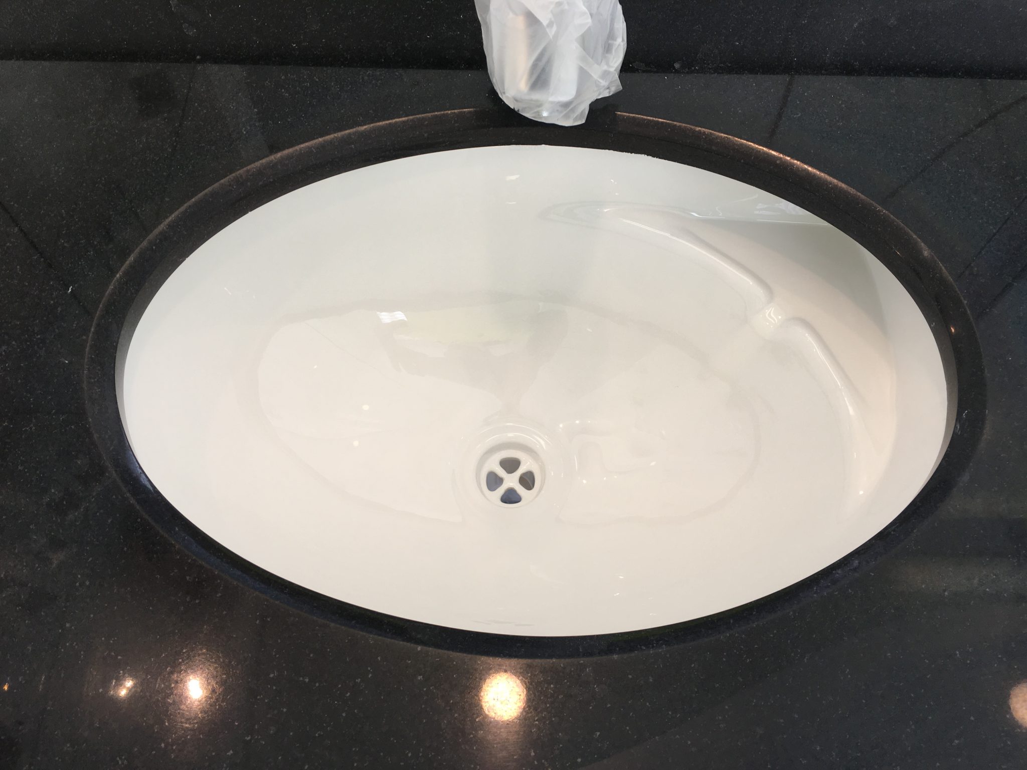 The image showcases an overhead view of an oval bathroom basin positioned atop a stylish black countertop. The basin's glossy white finish contrasts elegantly with the dark surface, highlighting a modern design ideal for contemporary bathroom renovations. A clear plastic bag, possibly used for protection during installation, is draped over the faucet area. This setting is indicative of a bathroom renovation project where new installations are being carefully managed and prepared for final touches. This aesthetic aligns well with Coastline Bath Resurfacing's commitment to providing stylish and functional bathroom renovations in Kyogle and its surrounding areas.