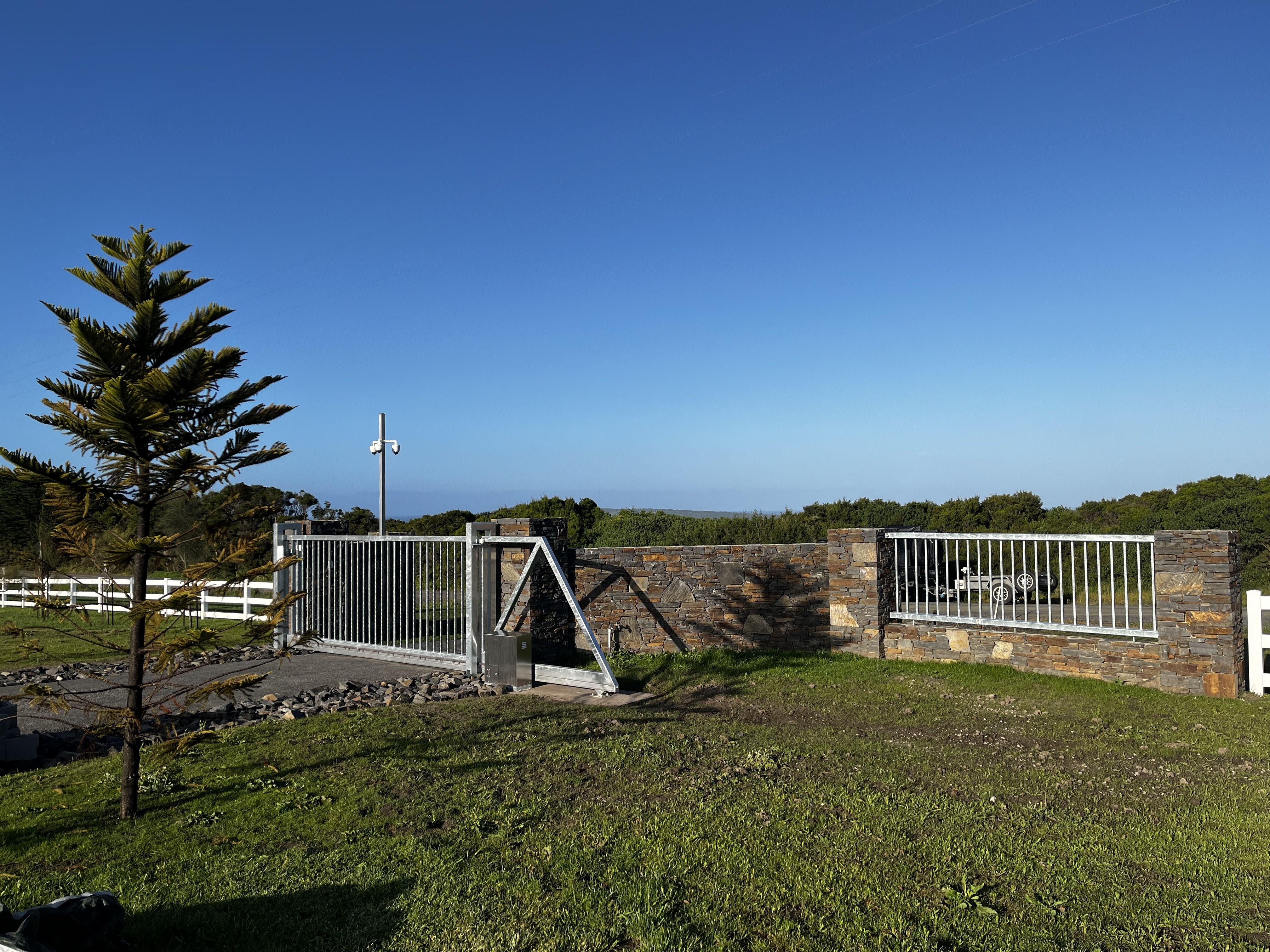 The image shows a modern entrance gate, accompanied by a stack stone wall, surrounded by well-maintained grass and a small tree in the foreground. The gate appears to be securely designed with a metal frame and sliding functionality, integrating aesthetics with security. The background features a clear blue sky and hints of greenery, indicating a tranquil environment, likely in a residential setting. This image emphasizes quality construction and attention to detail, reflecting the work ethos of Node Constructions.