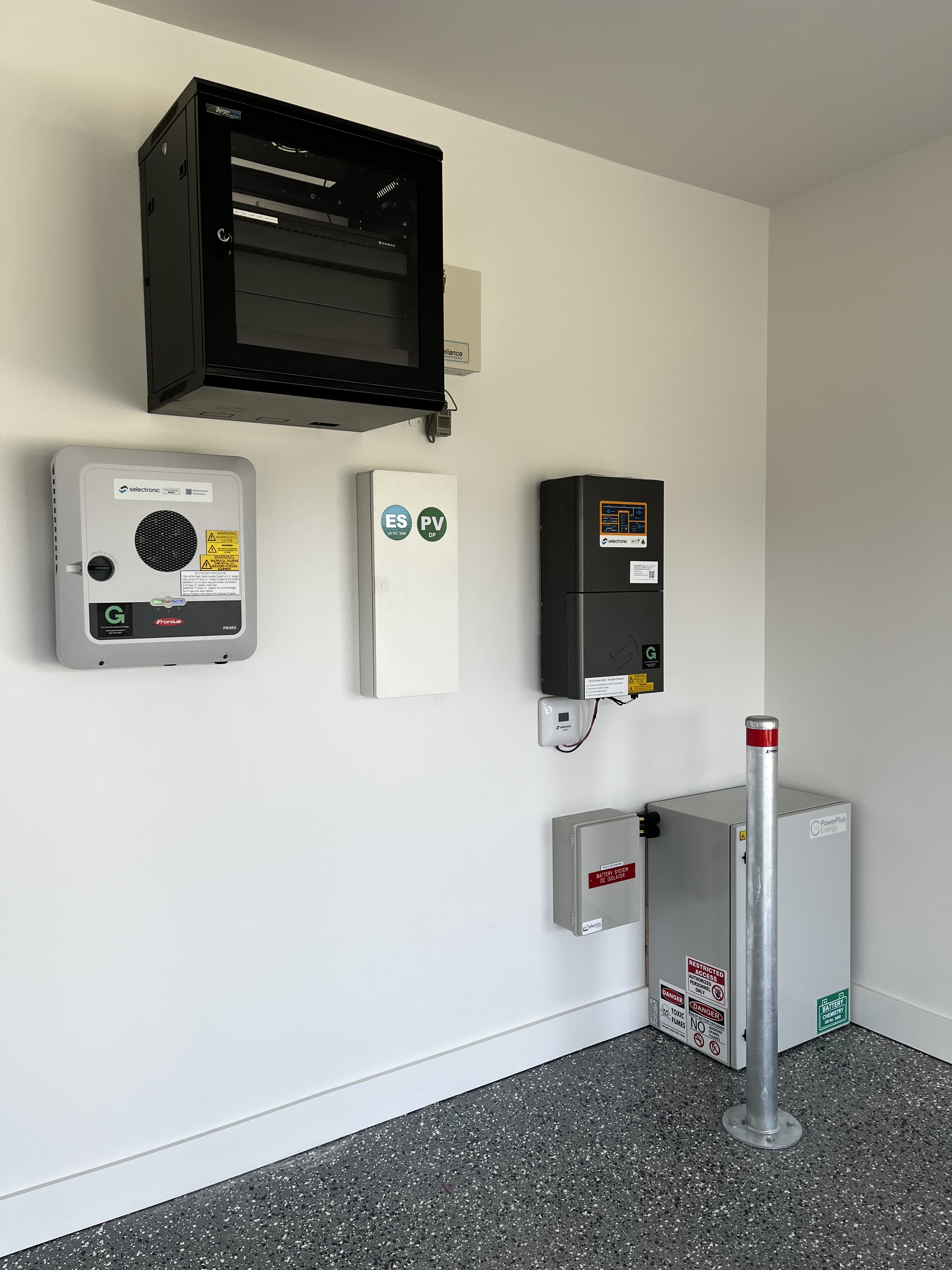 The image displays a wall-mounted installation featuring various electrical units, likely associated with a building project. Prominently, there is a black enclosure, possibly a control panel or a junction box, mounted higher on the wall. Below it, a grey inverter labelled 'G', essential for converting solar power, is visible alongside other units. These units may include a safety disconnect and monitoring systems, essential for energy management and compliance in contemporary building projects. This setup is typical in modern constructions, especially those integrating renewable energy sources like solar power.