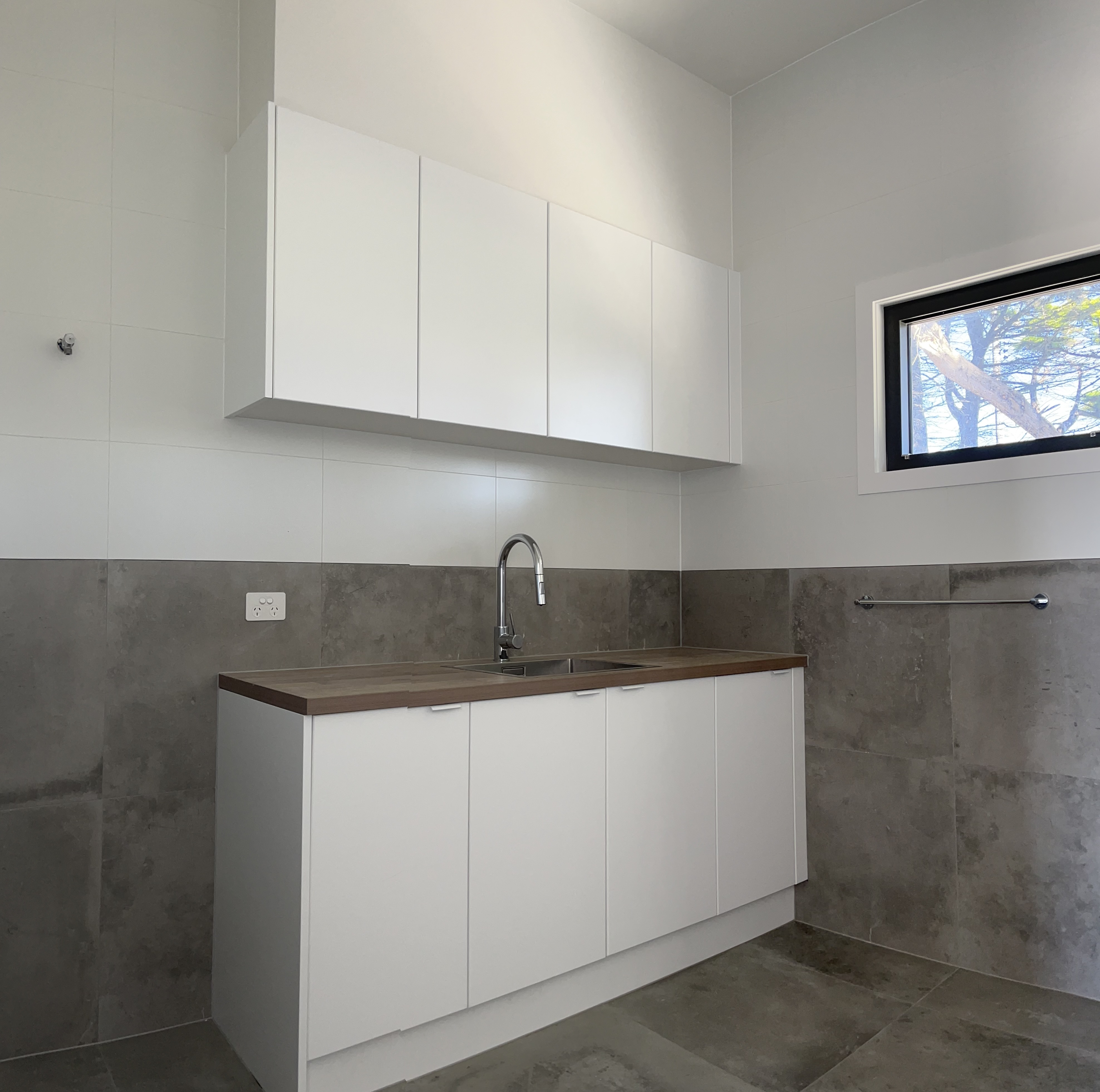 The image displays a modern kitchen area featuring sleek white cabinetry and a minimalist design. The upper cabinets are mounted on the wall, providing ample storage space, while the lower cabinets combine a white finish with a natural wood countertop. The space is well-lit and showcases a stylish stainless-steel sink with a contemporary tap. The wall behind the sink is finished with large grey tiles, complementing the overall aesthetic of the kitchen. This setting reflects a contemporary approach ideal for residential projects.