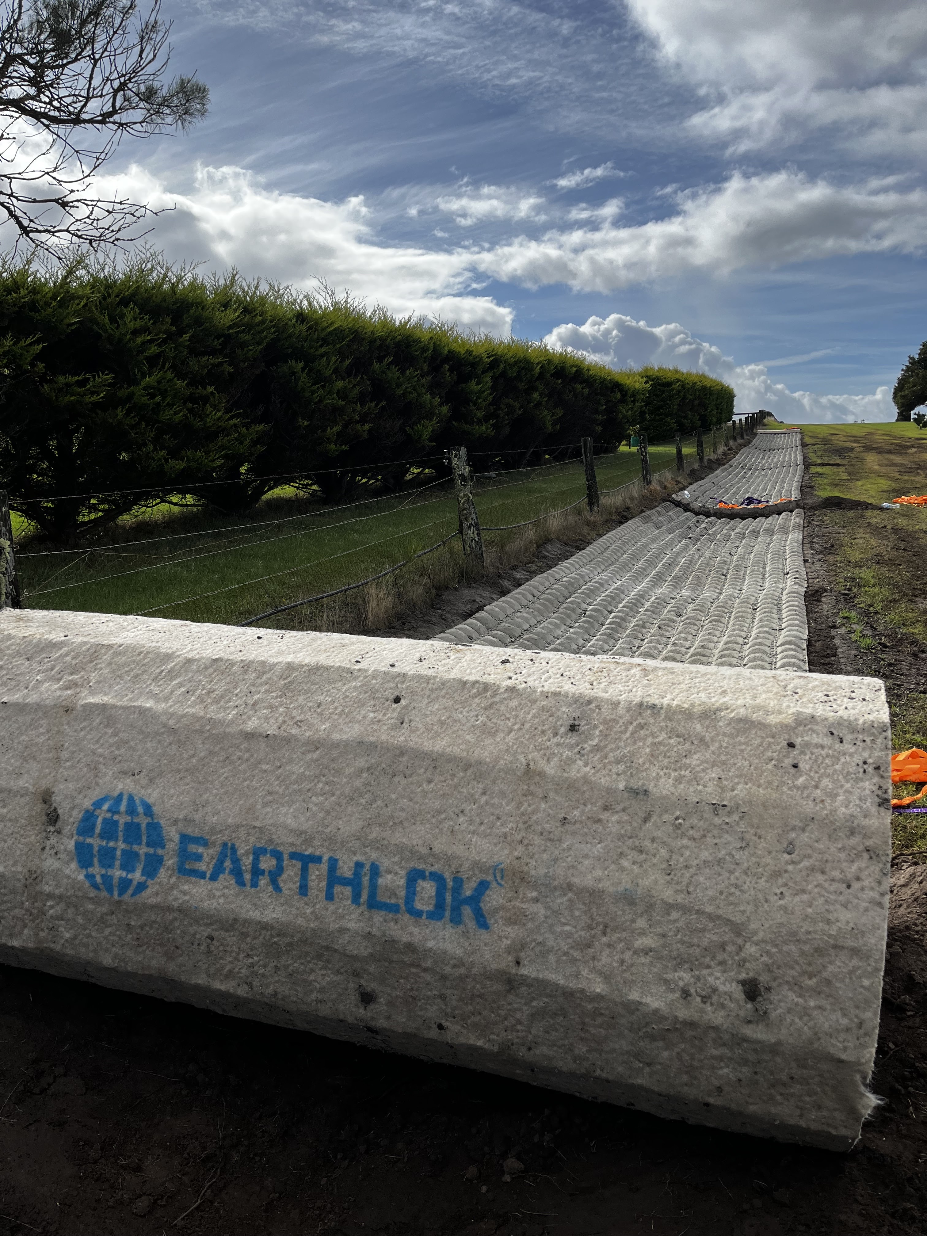 The image depicts a stretch of constructed surface, likely a pathway or access road, made from interlocking concrete blocks. These blocks are positioned on a prepared earth base, framed by green vegetation in the background, which suggests the site is in a semi-rural or suburban area. The sky is partly cloudy, indicating a good day for construction activity. A visible branding of 'EARTHLOK' on the concrete suggests it may be a product in use for stabilisation or drainage purposes. The setting implies that Node Constructions is engaged in developing durable and efficient access solutions, blending construction with environmental considerations.
