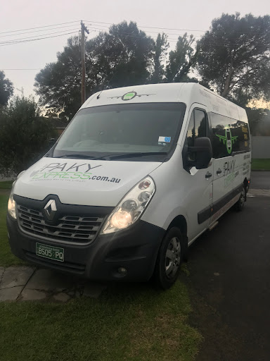 The image features a white Renault van, which is part of the Paky Express fleet, parked on a grassy area, possibly near an airport or public transport hub. The vehicle's exterior is branded with the Paky Express logo clearly visible on the side, indicating it provides airport shuttle services. The surroundings appear calm, likely early in the morning or late afternoon given the soft lighting conditions. The van is designed to accommodate passengers and their luggage, emphasising the convenience of their airport transfer services from areas like Hallam, Eumemmerring, and Endeavour Hills.