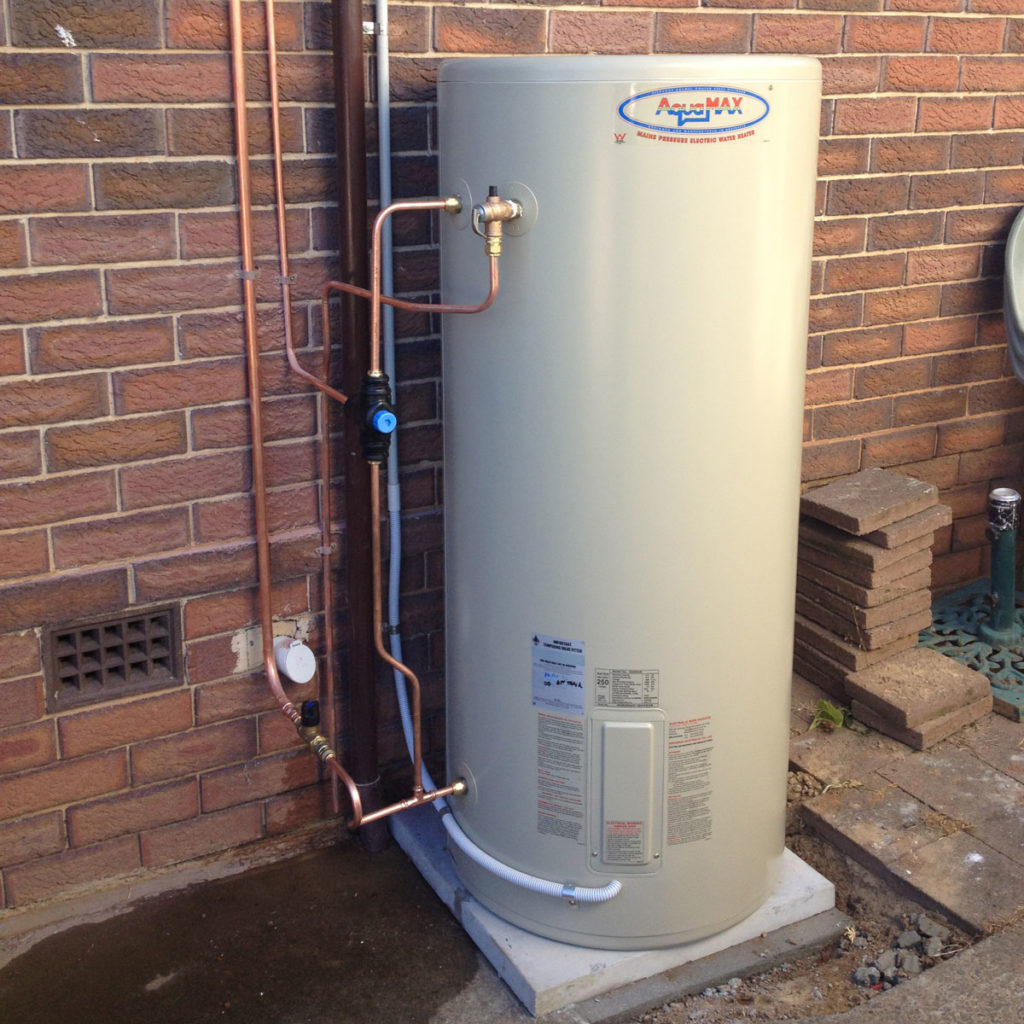 The image shows a hot water system, specifically an Aquamax water heater, installed outside against a brick wall. The heater features metal pipes for water inlet and outlet, which are connected with copper fittings. There’s a pressure relief valve and a water meter visible, indicating proper installation for safety and functionality. This type of appliance is crucial for electrical work in residential areas, emphasizing the importance of qualified electricians in the servicing of hot water systems.