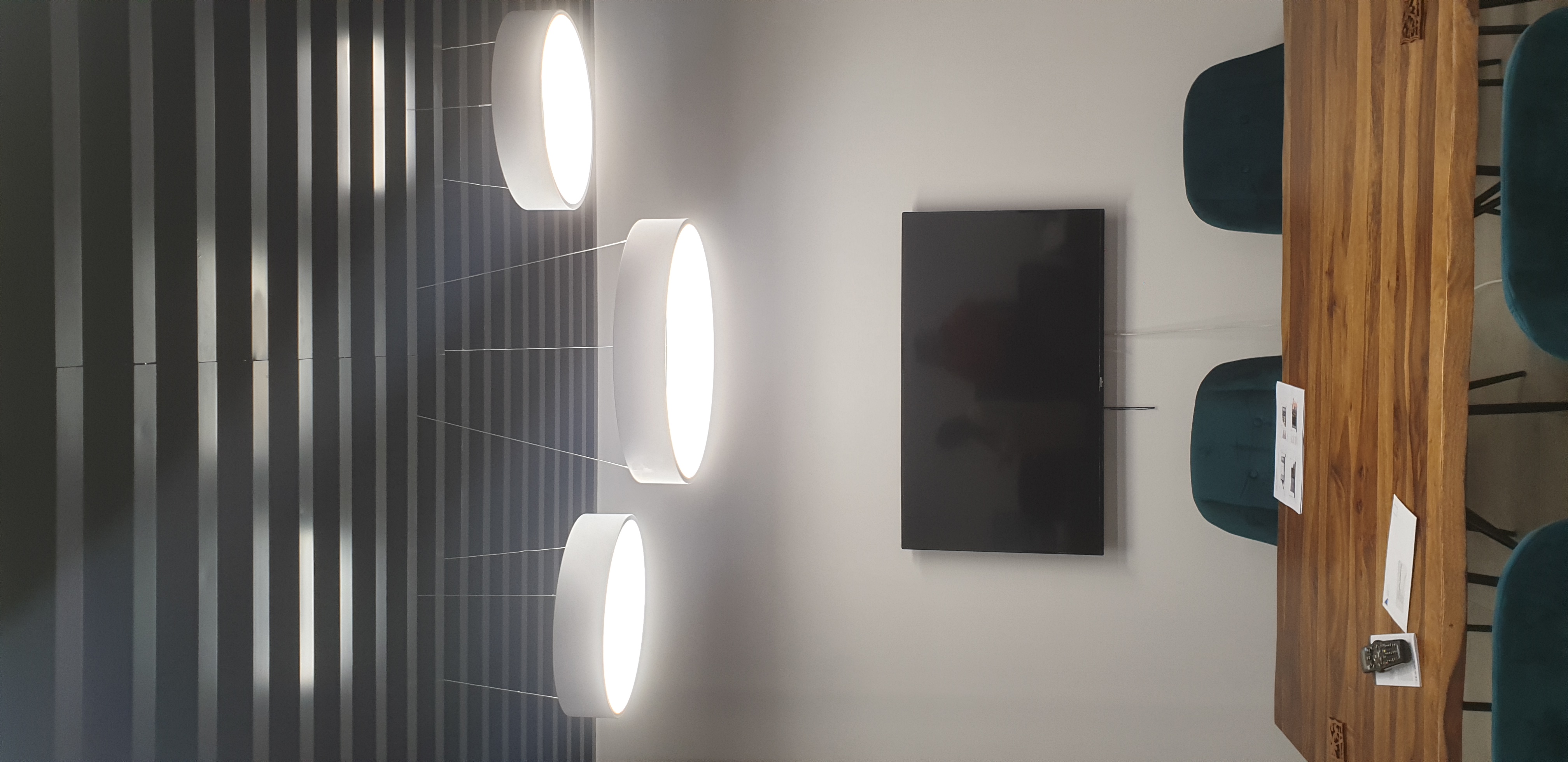 The image features a modern meeting room with a wooden table and three circular pendant lights hanging from a ceiling with horizontal metal strips. The lights emit a bright, diffused glow, enhancing the room’s ambience. On the wall opposite the table, there is a large flat-screen television, likely for presentations. Several papers are spread out on the table, suggesting preparation for a meeting or collaborative work. The overall design conveys a professional and contemporary atmosphere suitable for business discussions.