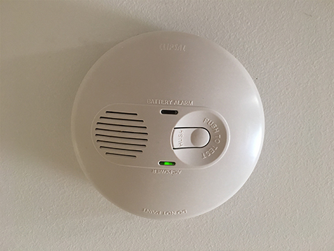 The image depicts a battery-operated smoke alarm or carbon monoxide detector mounted on a ceiling. The unit is circular with a smooth white finish, featuring a grill for sound emission and a test button. There is a green indicator light that suggests the device is functioning properly. This is a crucial safety device for any home or business, especially for electrical installations and safety compliance.