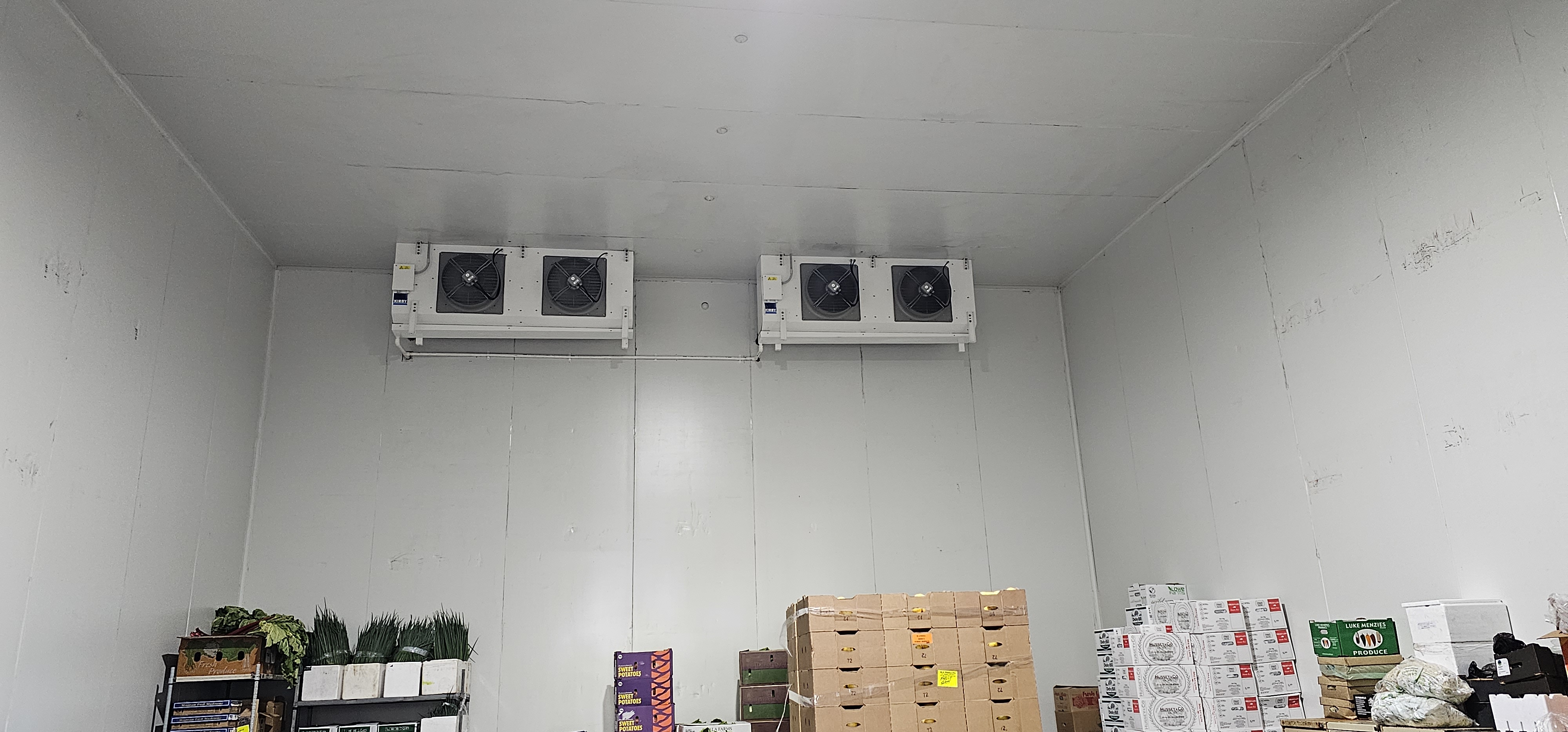 The image shows a commercial refrigeration room with two large wall-mounted cooling units installed near the ceiling. The room is a cold storage space with white insulated walls, designed to keep perishable goods fresh. Inside the room, there are stacks of cardboard boxes and containers, some of which are labelled with produce names like sweet potatoes. Various fresh vegetables are visible, indicating this refrigerated space is used for storing fresh produce. The cooling units are industrial-grade, appropriate for commercial refrigeration needs in businesses such as grocery stores or markets.