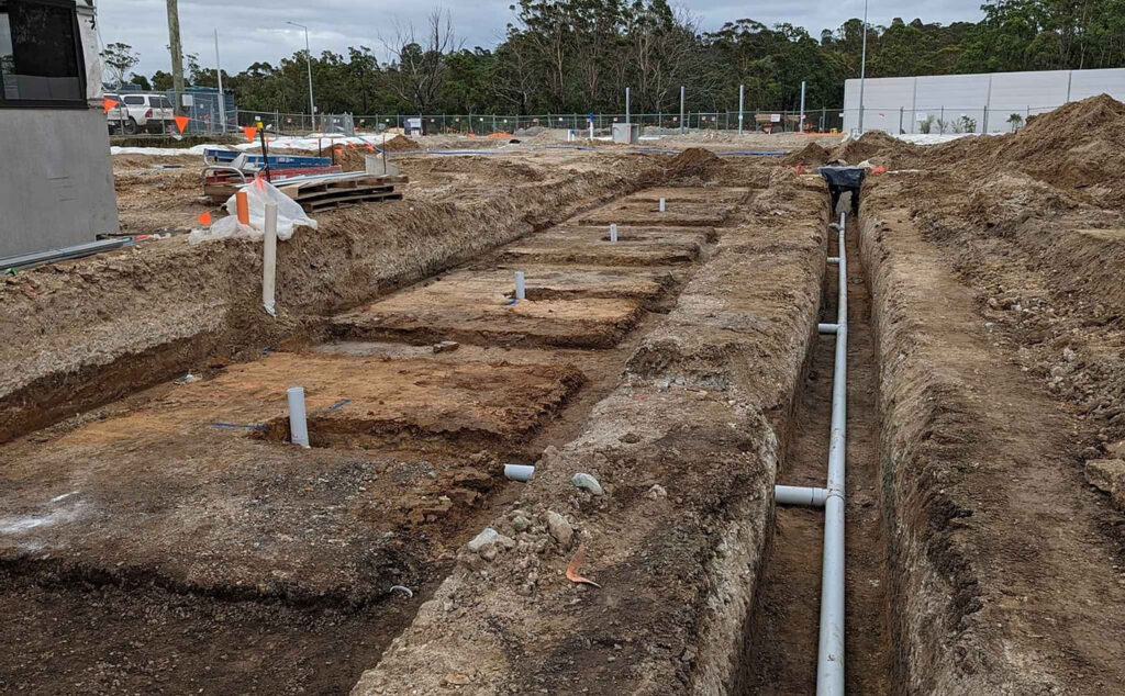 The image depicts an excavated area at a construction site, showcasing trenches dug into the earth. Within the trenches, white PVC pipes are visible, likely indicating newly installed plumbing or drainage systems. The surroundings include disturbed earth and construction materials, suggesting ongoing building work. This installation may be part of a foundational plumbing system for a new structure, crucial for waste disposal and water supply. The site appears to be in a developing area, possibly in or near Maroochydore.
