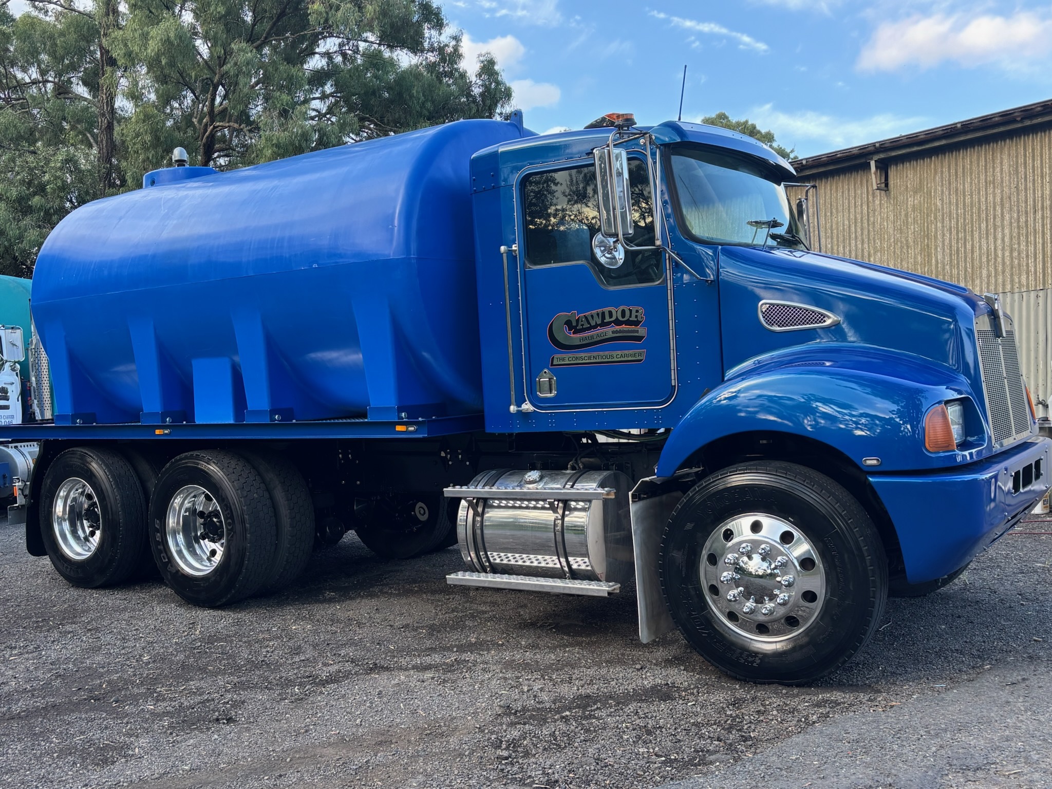 The image features a striking blue truck, specifically a tanker vehicle, parked prominently on a gravel surface. The truck has a robust and shiny design, indicative of its heavy-duty capabilities. Its large tank suggests it is likely used for transporting liquid or bulk materials, essential for a transportation service. The background shows a structure, possibly a depot or garage related to the Cawdor Haulage business, indicating that this vehicle is part of their fleet operations. The presence of the truck underscores the company's commitment to providing reliable transport services in the Cawdor and Razorback areas, supporting various logistical needs within these regions.