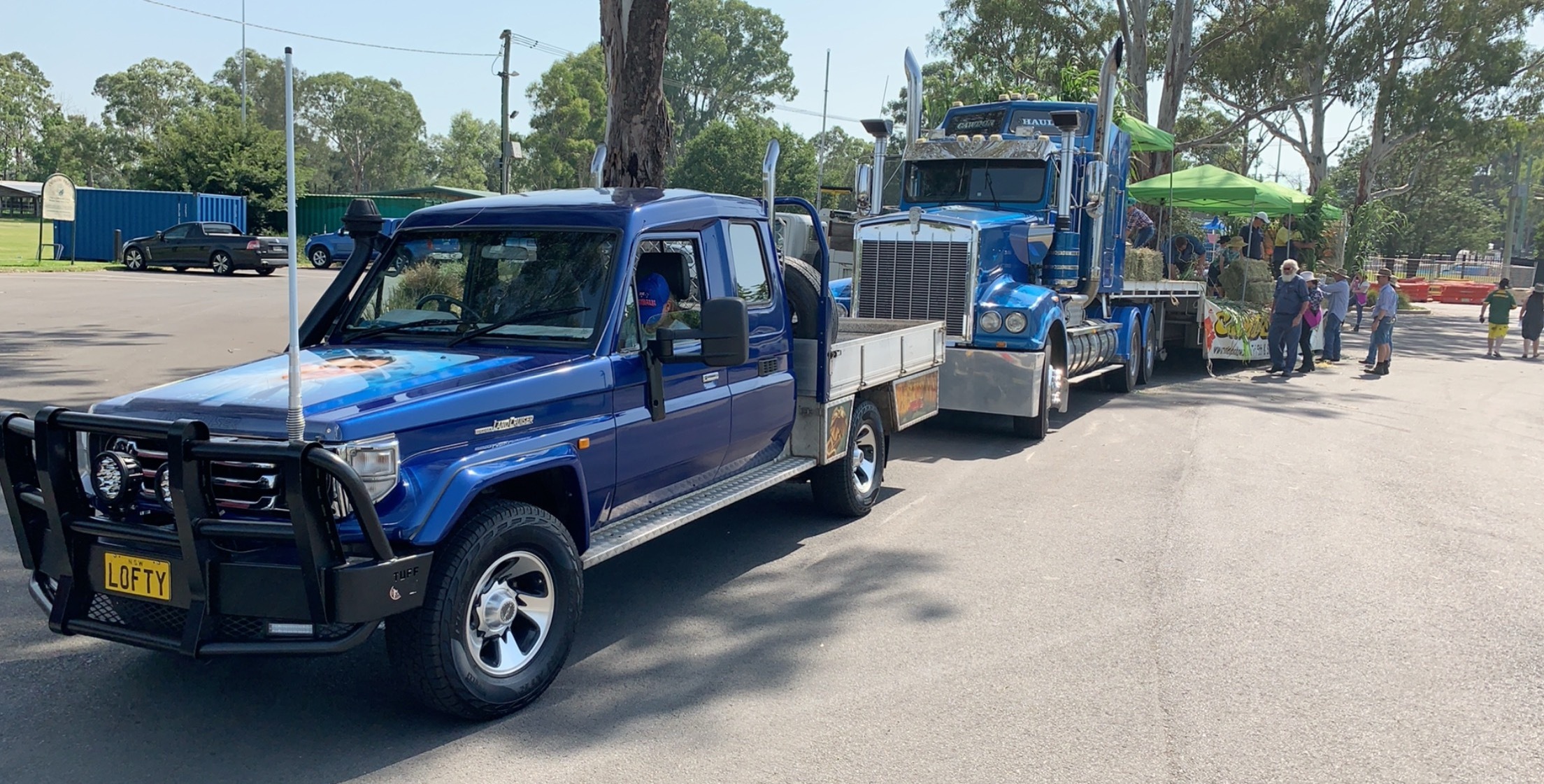 The image showcases a transport service setting with two distinct vehicles parked along a wide road, framed by trees. The vehicle in the foreground is a blue utility truck, likely part of Cawdor Haulage's fleet, exhibiting robust features suitable for cargo transport. The truck has a tray at the back, perfect for carrying heavy loads, and displays the business's name. Behind it, there's a larger blue truck, possibly a prime mover, indicating Cawdor Haulage's capacity to handle extensive transport needs. Both vehicles are stationed in a setting that suggests a busy day of operations, possibly waiting for clients to load or unload goods. Nearby, the presence of tents and people signifies an event or community activity, highlighting the transport service’s engagement with the local community in Razorback, NSW.