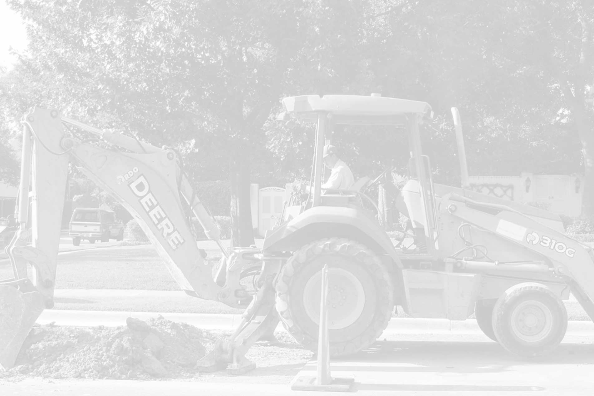 The image shows a John Deere 310G backhoe loader in operation. It is actively digging into the ground, moving earth, indicative of earthmoving and excavation work. A person is seated in the operator's cabin controlling the machinery. The background includes trees and a residential area, suggesting that the work might be part of a construction or landscaping project within a community or residential zone. This scene represents the heavy equipment used by an earthmoving contractor like Skip Hire Phillip Island to carry out earthmoving and excavation jobs.
