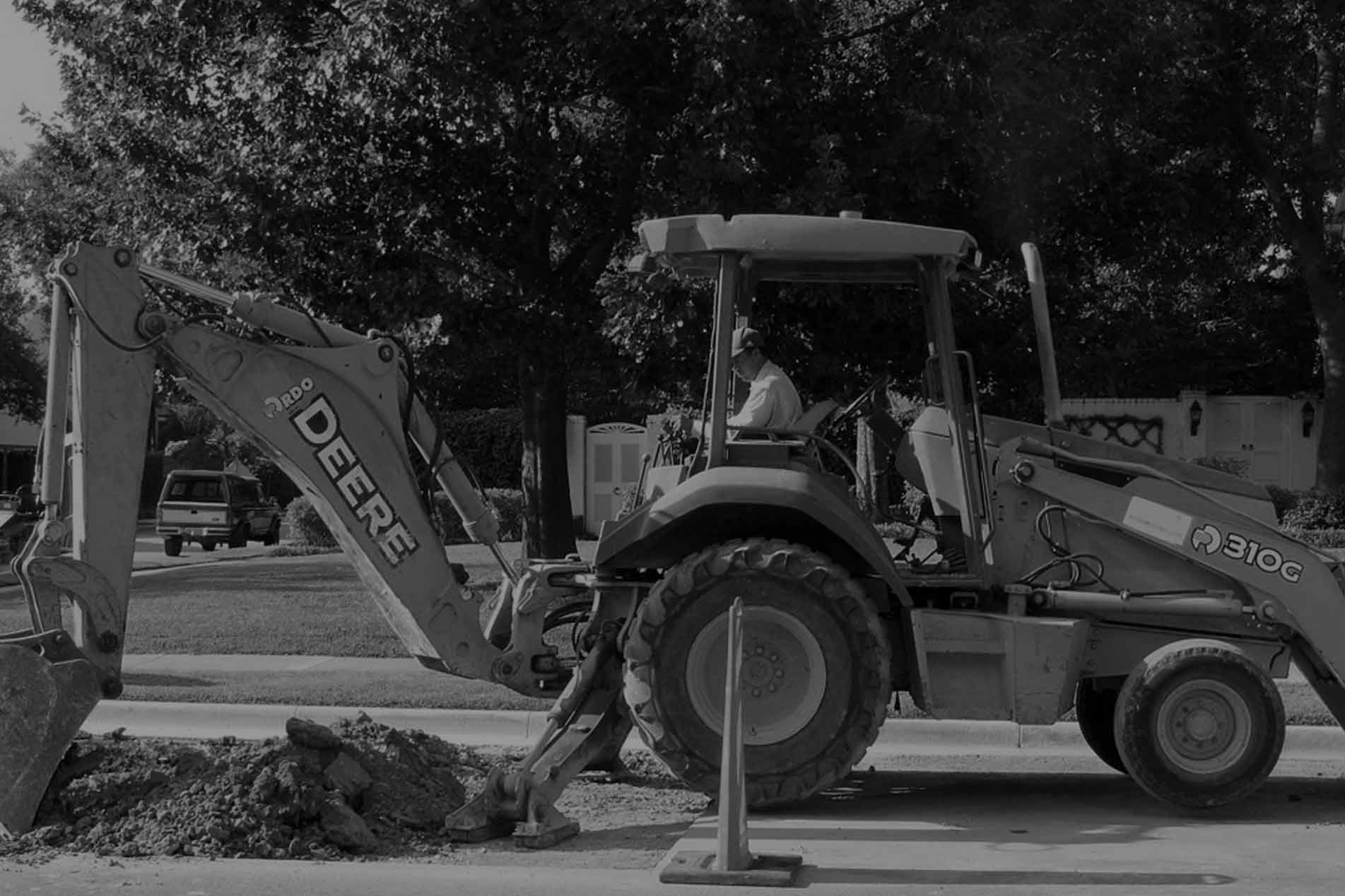 The image shows a John Deere 310G backhoe loader actively digging into the soil on a paved road. The machine is operated by a person seated inside the cabin, focused on the earthmoving task. The surroundings include trees and residential features, indicating the earthmoving work is being carried out in a suburban or semi-rural area. A pile of excavated soil is visible next to the digging site, showcasing typical earthmoving activity that is integral to construction or landscaping projects.