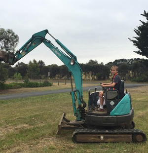 A young person is operating a small blue-green tracked excavator in a grassy field. The excavator is a compact earthmoving machine used for digging and landscaping. The background shows a rural setting with trees and an overcast sky, typical for earthmoving operations in Smiths Beach and surrounding areas. This image represents earthmoving services suitable for residential or light commercial projects.