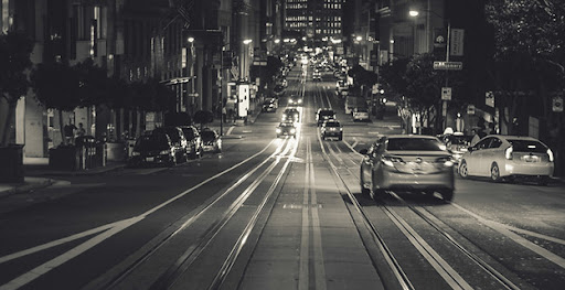 The image depicts a quiet street scene at night, with illuminated buildings lining the street. A few vehicles are visible on the road, and the faint glow of streetlights casts a soft light over the area. The track of tram lines can be seen running down the centre of the street, highlighting its urban setting. The overall ambience is calm, with an emphasis on the peacefulness of the city at night, which could reflect a potential customer’s journey to a mechanic service like True Blue Mechanical in Wynnum.