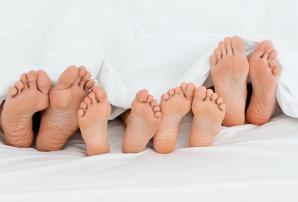 The image showcases a variety of feet, including both adult and children's feet, arranged side by side on a white bed cover. This visual represents the theme of family and care, highlighting the importance of foot health across different age groups, which is relevant to a podiatry practice. The feet are well-groomed and indicative of ongoing foot care, suggesting a focus on the health and well-being of the entire family. The setting implies comfort and homeliness, aligning with the values of BAY PODIATRY CLINIC in providing a welcoming environment for patients of all ages.