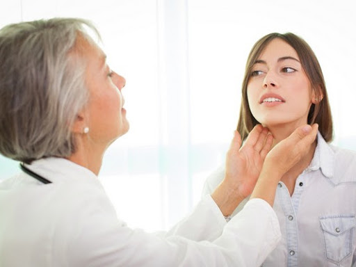A medical professional, likely a doctor, is examining a young female patient's neck, possibly checking for lymph nodes or thyroid issues, within a well-lit clinical setting. This interaction depicts a thorough health consultation at a medical centre.