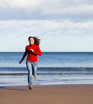 A young woman in a red sweater and jeans is joyfully running along the beach near the ocean under a partly cloudy sky. The beach setting suggests a healthy, active lifestyle, which aligns with the wellness focus of Dr Nadia Tejani's Medical Centre in Maroubra, NSW.