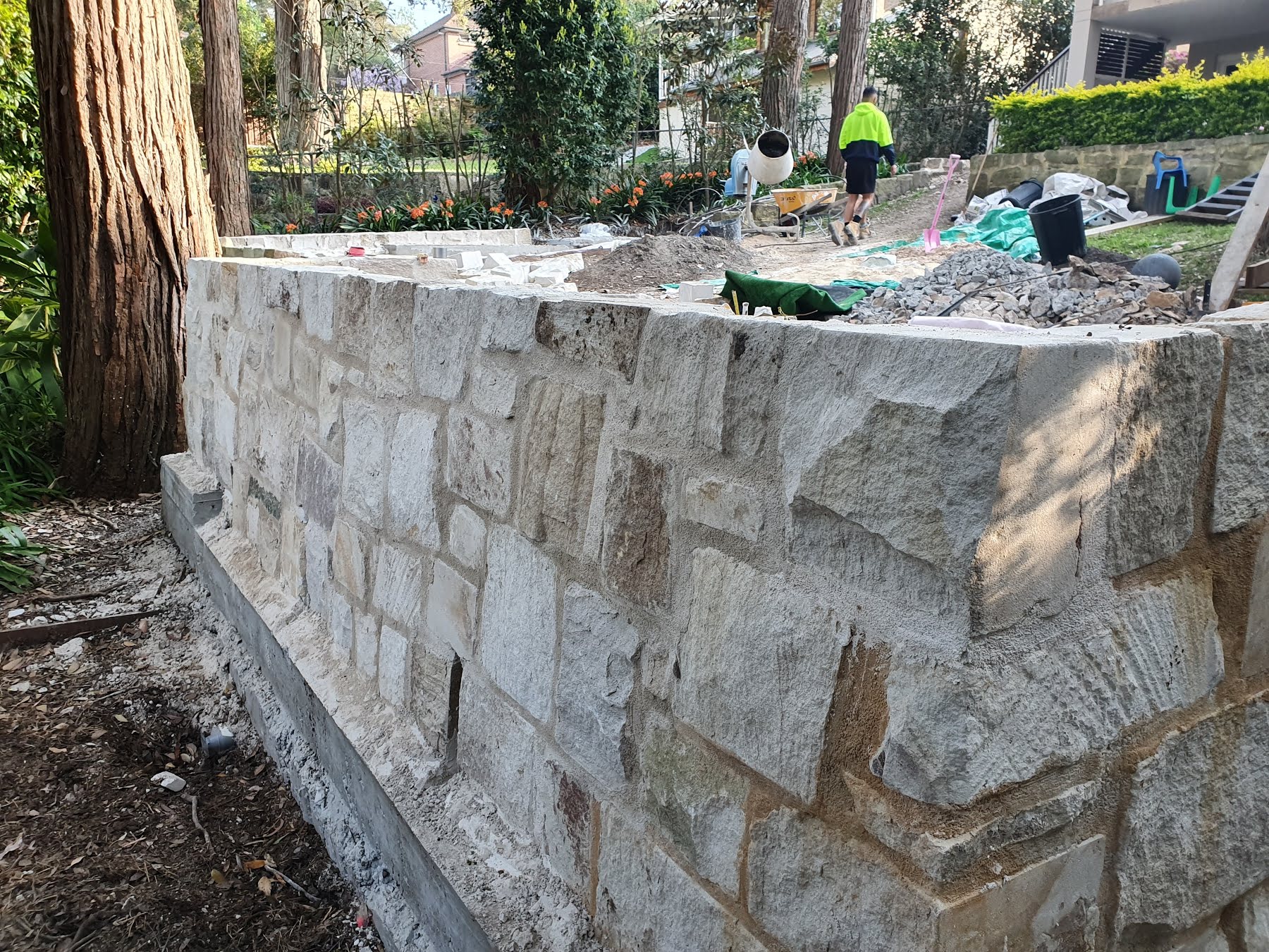 The image showcases a partially constructed stone wall, indicative of masonry work in progress. The wall features various sizes and shapes of stone blocks, carefully arranged, highlighting the skilled craftsmanship of the bricklayer. In the background, a worker dressed in bright yellow attire is visible, engaged in tasks related to the ongoing project, likely ensuring the site is prepared for the next steps. The surrounding area is cluttered with tools and materials related to bricklaying, such as a cement mixer and other masonry equipment. This scene exemplifies the quality of work that Masters Bricklaying and Stonemasonry delivers to clients in Cremorne and surrounding suburbs like Mosman and Cammeray.