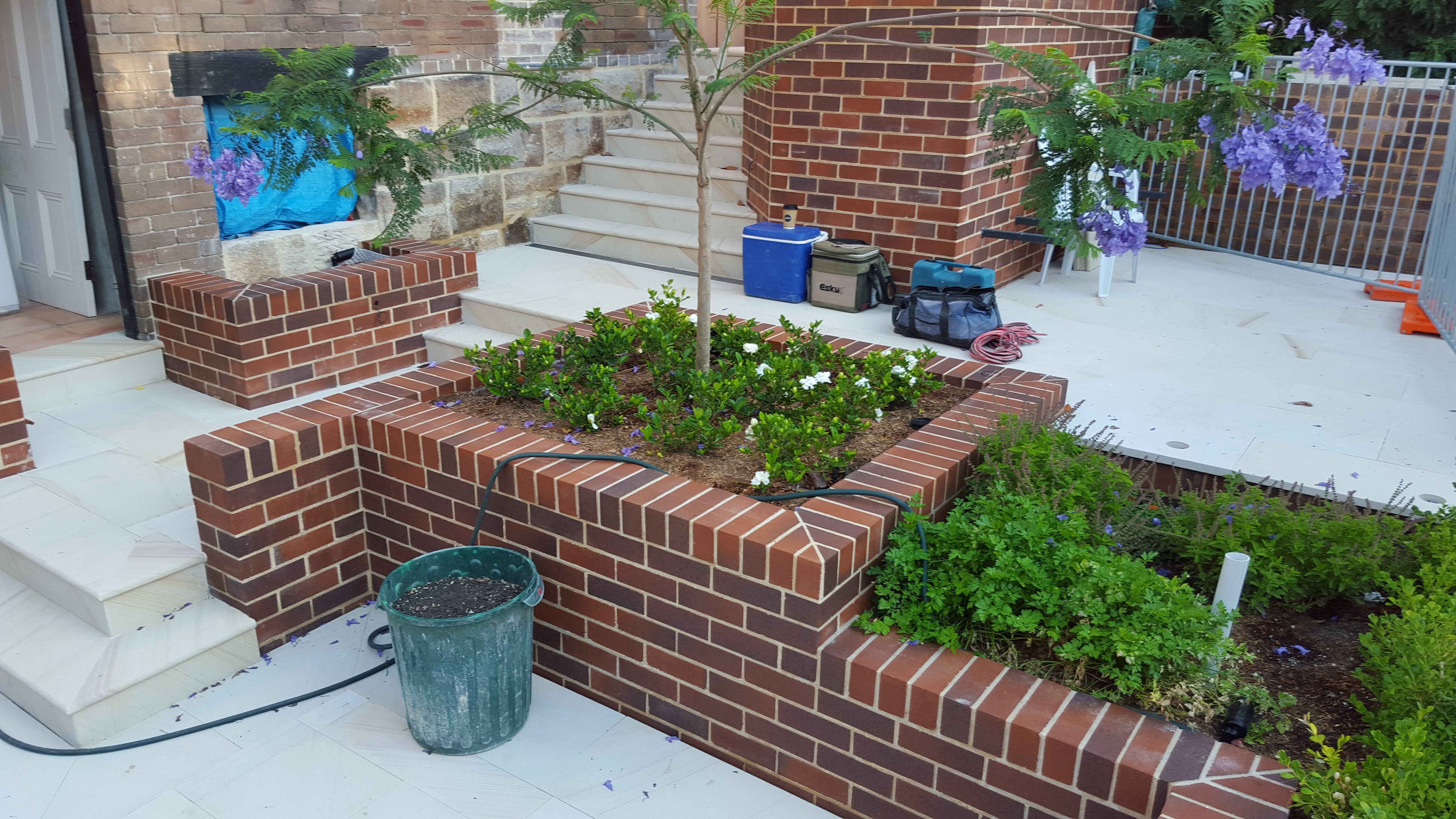 The image showcases a beautifully constructed brick garden bed as part of a landscaping project completed by Masters Bricklaying and Stonemasonry. The brickwork is neatly laid, displaying a consistent pattern and quality craftsmanship inherent to bricklaying. Surrounding the central garden area filled with plants and decorative flowers, the brick border enhances both aesthetics and functionality. Adjacent to the garden bed, stone steps lead to the entrance of a home, indicating that this installation is part of a larger outdoor renovation. Tools and equipment can also be seen nearby, suggesting ongoing landscaping work. With the combination of traditional bricklaying techniques and modern landscaping elements, this project highlights the expertise and service quality of Masters Bricklaying.