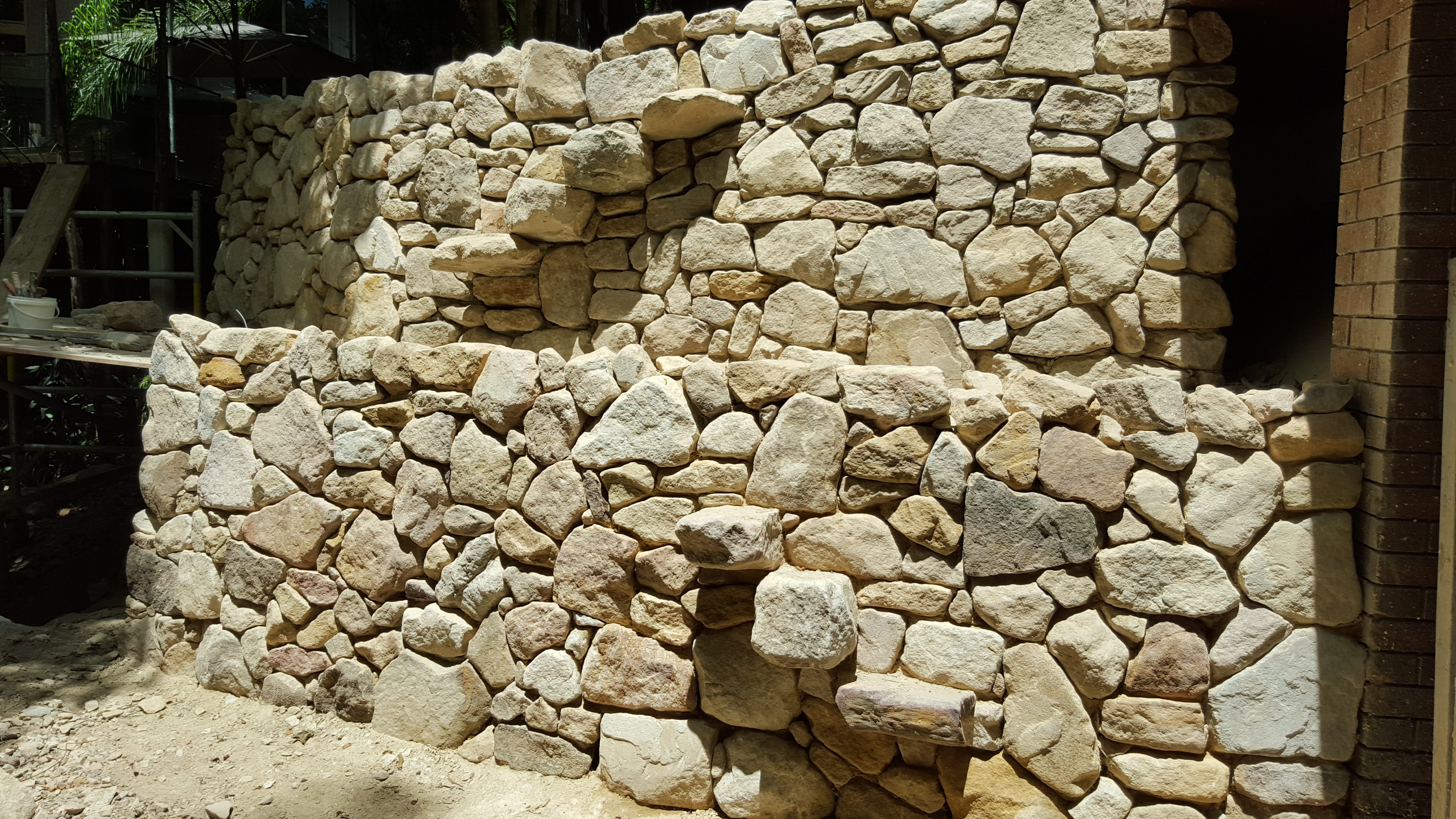 The image showcases a newly constructed stone wall, demonstrating skilled craftsmanship common in bricklaying and stonemasonry. The wall is composed of various sizes of light-coloured, rough-cut stones arranged intricately to create a natural yet structured appearance. There are visible gaps between some stones, characteristic of a dry-stone wall method, which allows for flexibility and drainage. The work reflects a strong understanding of aesthetics and durability, ideal for outdoor landscaping or property boundaries in residential areas. The surrounding site shows evidence of ongoing construction, indicating that this bricklayer is actively working on projects in Cremorne, enhancing the area's architectural appeal.